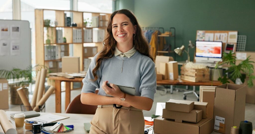 A small business owner smiles confidently in her workspace, surrounded by stock and packaging materials.