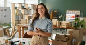 A small business owner smiles confidently in her workspace, surrounded by stock and packaging materials.