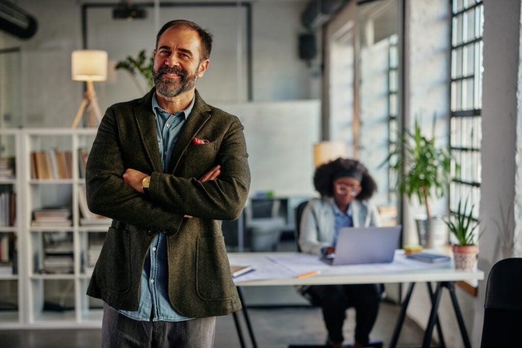 A confident South African business owner stands with arms crossed in a modern office, with a colleague working at a laptop in the background.